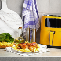 Fritadeira elétrica amarela sobre bancada cinza ao lado de prato com frango assado e batatas, tábua com alface, tomate, limão e azeite ao fundo.