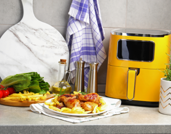 Fritadeira elétrica amarela sobre bancada cinza ao lado de prato com frango assado e batatas, tábua com alface, tomate, limão e azeite ao fundo.