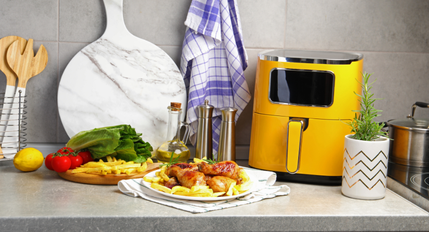 Fritadeira elétrica amarela sobre bancada cinza ao lado de prato com frango assado e batatas, tábua com alface, tomate, limão e azeite ao fundo.