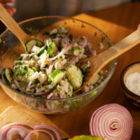 Salada com pepino fatiado, cebola roxa e ervas verdes sendo misturada com utensílios de madeira em uma tigela de vidro; ao lado, aparecem sal, alho e pimentas verdes.