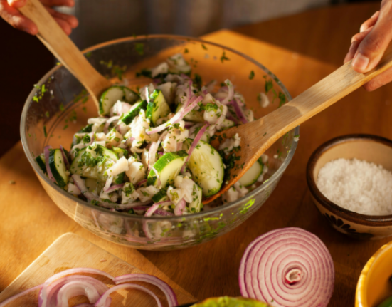 Salada com pepino fatiado, cebola roxa e ervas verdes sendo misturada com utensílios de madeira em uma tigela de vidro; ao lado, aparecem sal, alho e pimentas verdes.