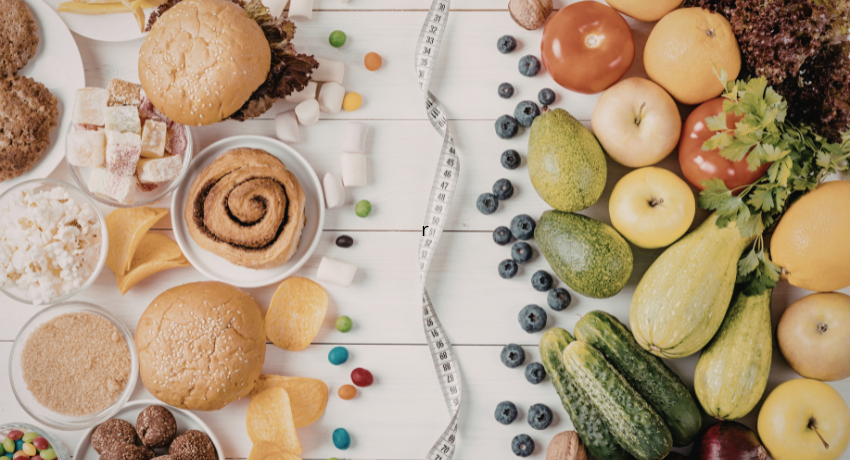 Comparação entre alimentos ultraprocessados, como hambúrguer e doces, e alimentos naturais, como frutas e vegetais, divididos por fita métrica sobre mesa branca.